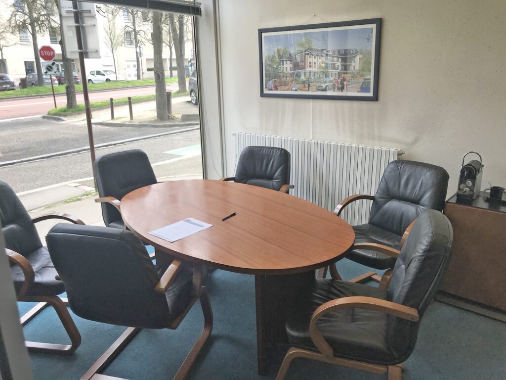 Une petite salle de conférence avec six chaises noires autour d'une table ovale en bois, avec un papier et un stylo sur la table, et une grande fenêtre montrant la rue à l'extérieur.