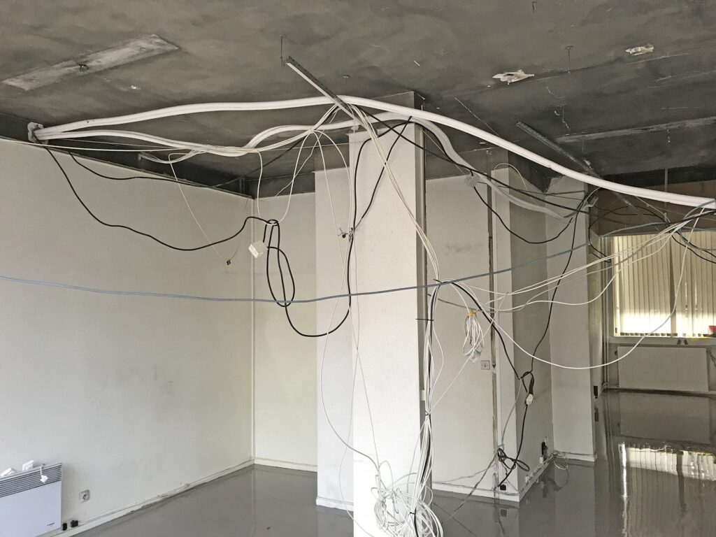 Des fils et des câbles électriques apparents pendent du plafond et des murs dans une pièce inachevée ou abandonnée, au béton nu et au sol réfléchissant.
