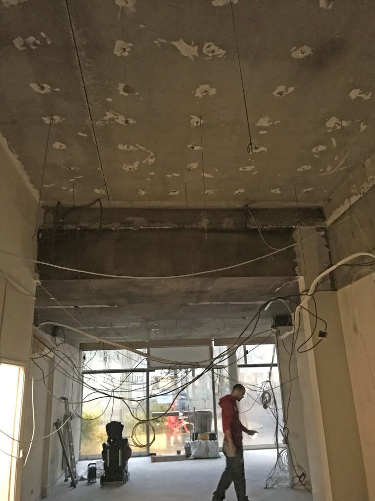 Un chantier de construction avec des fils apparents qui pendent du plafond, des murs inachevés et une personne qui marche dans la zone.