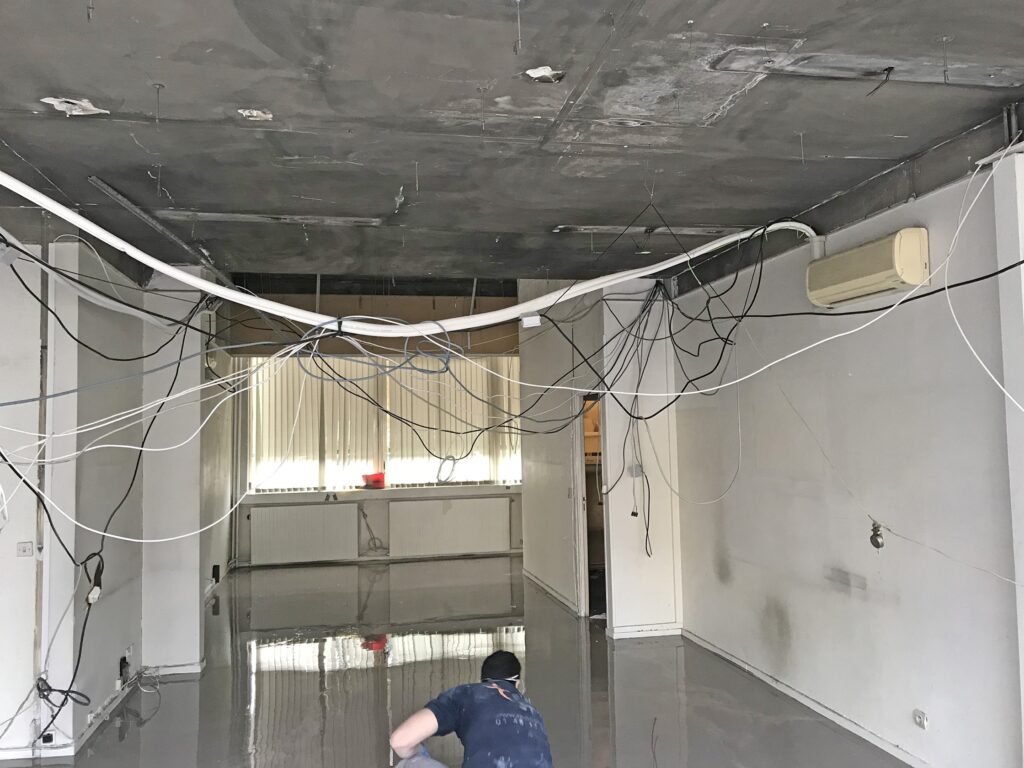 Une personne est agenouillée sur le sol brillant d'une pièce en cours de rénovation, où des câbles apparents pendent du plafond inachevé.