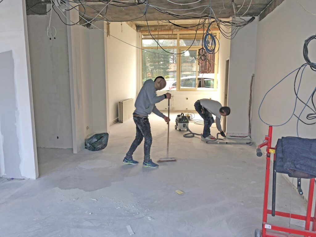 Dans une pièce partiellement rénovée, deux personnes nettoient les débris de construction ; des fils électriques à nu pendent du plafond et des murs inachevés sont visibles.