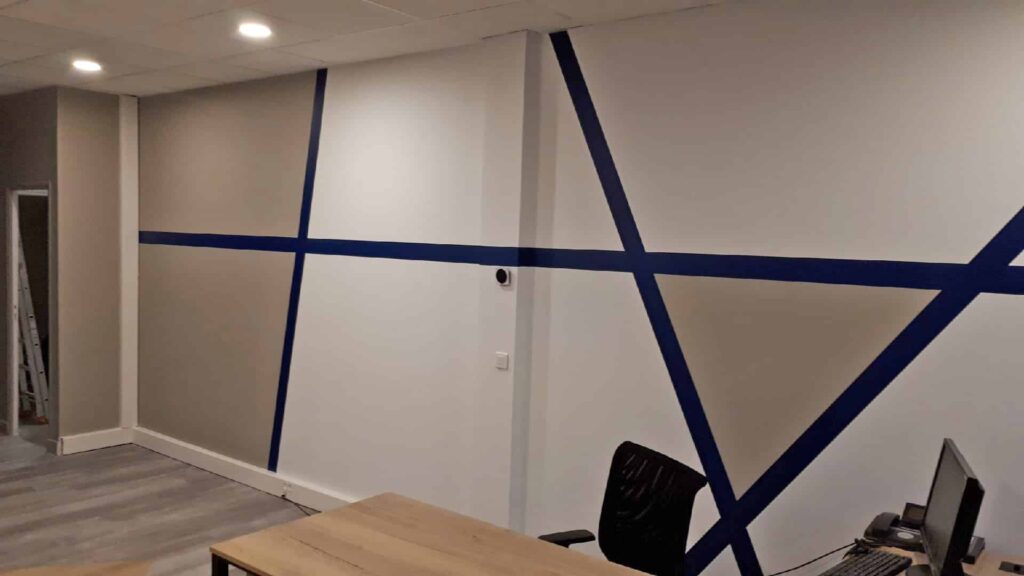 Mur de bureau avec de grandes lignes géométriques de ruban adhésif bleu traversant des sections peintes en blanc et en beige ; bureaux et chaises au premier plan, plafonniers au-dessus de la tête.