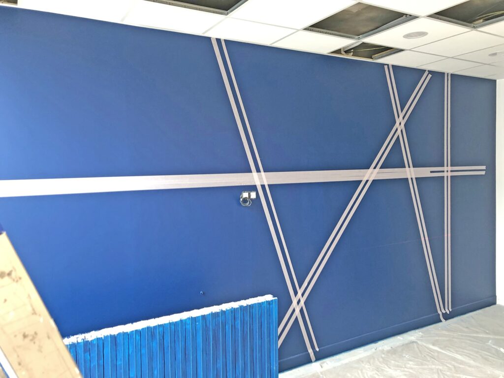Un mur bleu avec des accents diagonaux et horizontaux argentés, sous un plafond partiellement achevé avec des panneaux apparents et quelques matériaux de construction visibles.