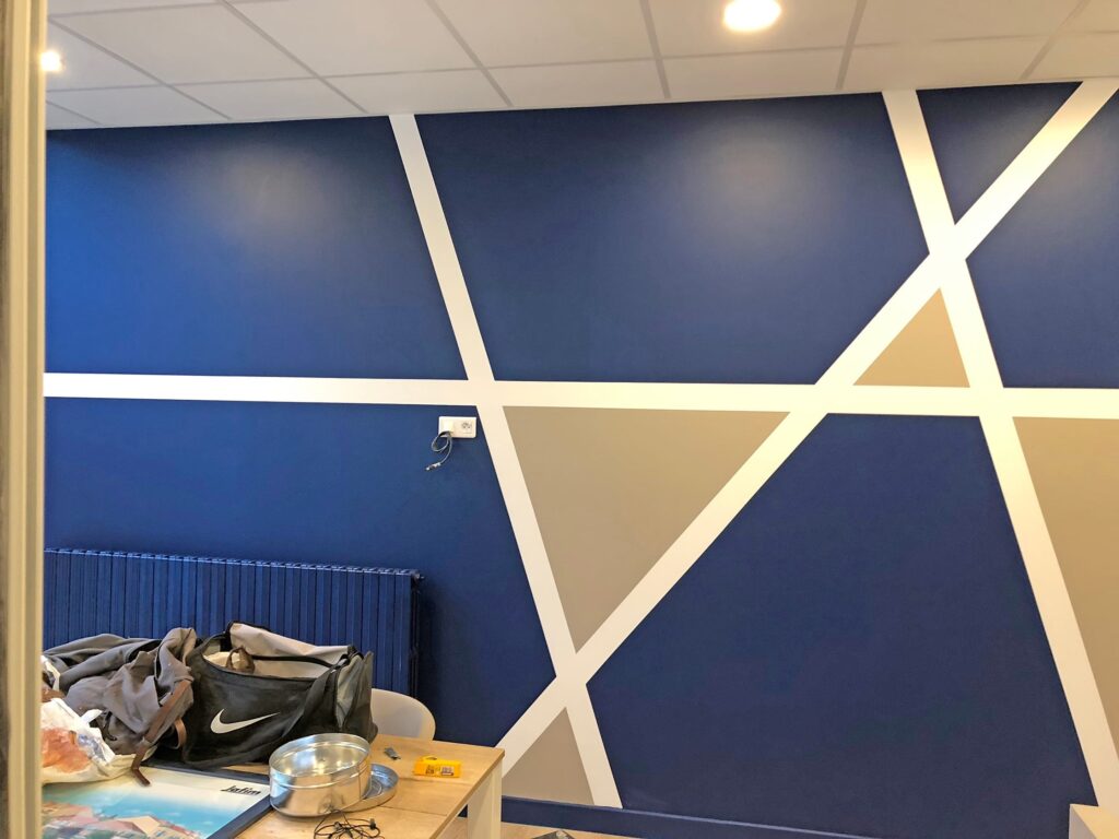 Mur peint de formes géométriques bleues, blanches et beiges ; plafonniers, radiateur et table encombrée visibles au premier plan.