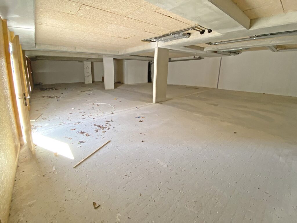 Un grand sous-sol vide et inachevé, avec un sol et un plafond en béton, des tuyaux apparents, des débris épars et la lumière du soleil qui pénètre par une fenêtre.