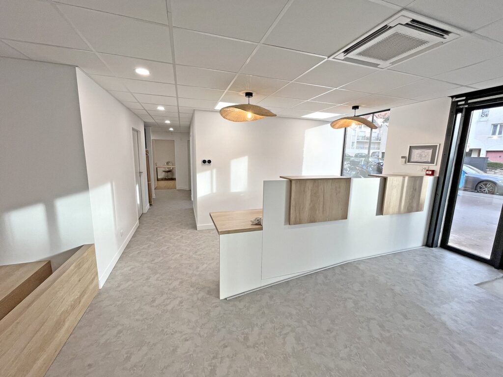 Espace de réception avec un bureau moderne en bois et blanc, des luminaires suspendus et un couloir menant à d'autres pièces ; de grandes fenêtres laissent entrer la lumière naturelle.