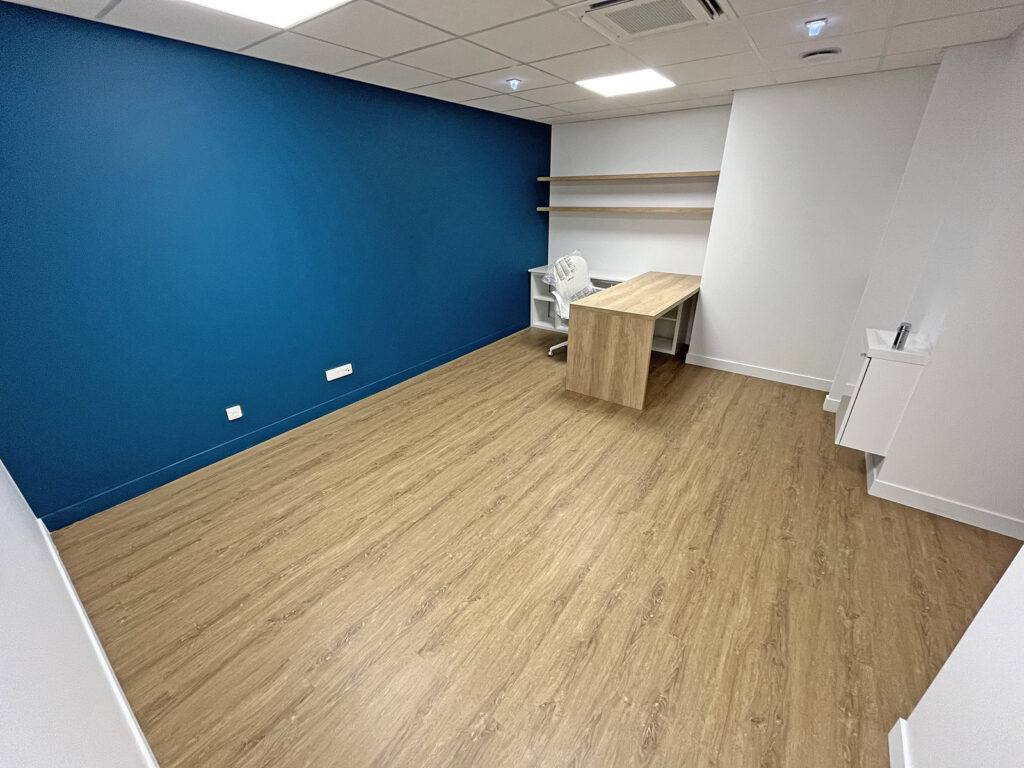 Bureau vide avec un sol en bois, un mur d'accent bleu, un bureau, une chaise de bureau blanche, des étagères flottantes et une petite armoire blanche.