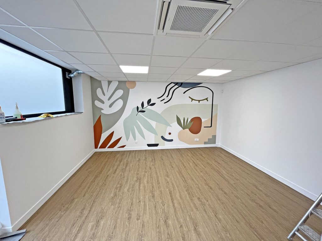 Une pièce moderne et vide avec un sol en bois, des dalles de plafond blanches et un mur orné d'une peinture murale abstraite colorée avec des formes géométriques et végétales.