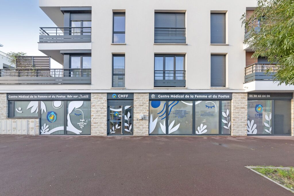 Vue de la rue d'une clinique médicale moderne avec de grandes fenêtres en verre affichant le nom "Centre Médical de la Femme et du Foetus" et des motifs décoratifs de feuilles blanches.