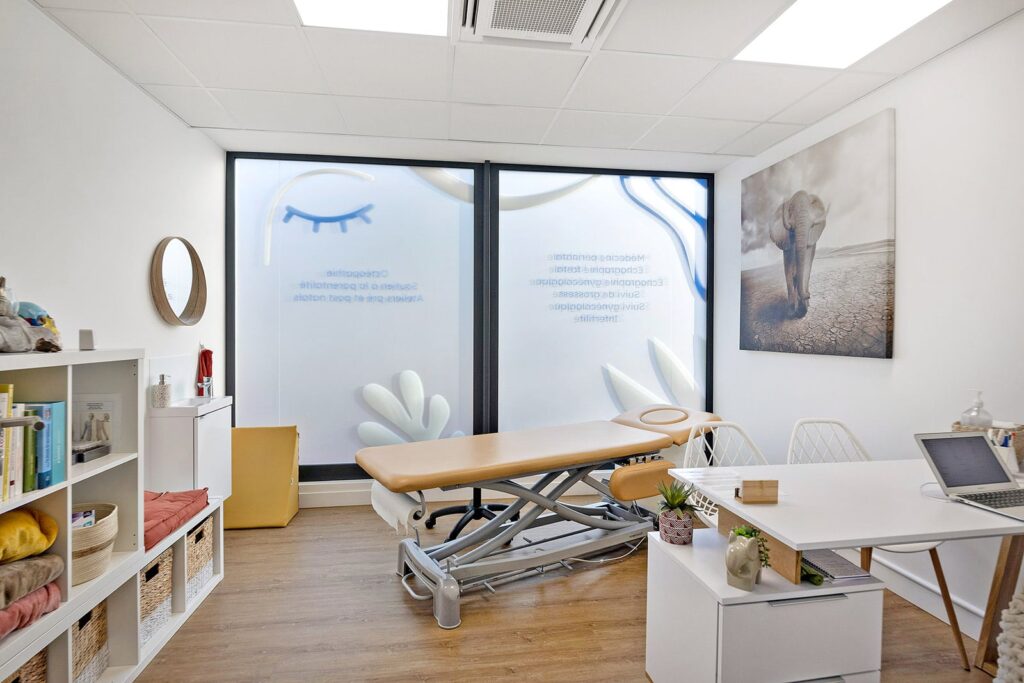 Une salle médicale ou thérapeutique propre et moderne avec une table d'examen, un bureau, un ordinateur, des étagères et un tableau mural représentant un éléphant. De grandes fenêtres givrées laissent entrer la lumière naturelle.
