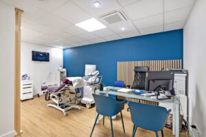 Une salle d'examen médical moderne avec un fauteuil d'examen, un appareil à ultrasons, un bureau avec du matériel de bureau, des chaises bleues et une télévision au mur.