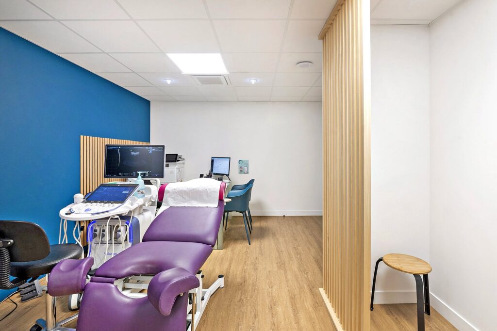 Une salle d'examen médical avec une chaise d'examen violette, un équipement d'échographie, un poste de travail informatique, un sol en bois, un mur d'accent bleu et une cloison de séparation en bois.