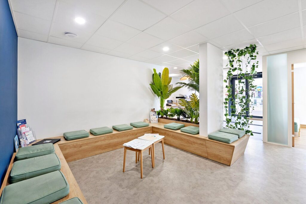 Une salle d'attente moderne avec des bancs en bois, des coussins verts, une petite table avec des magazines, des plantes d'intérieur et un éclairage naturel lumineux.