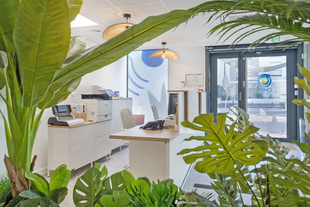 Un espace de réception moderne avec des bureaux, des équipements de bureau et de grandes plantes vertes près d'une porte d'entrée en verre.