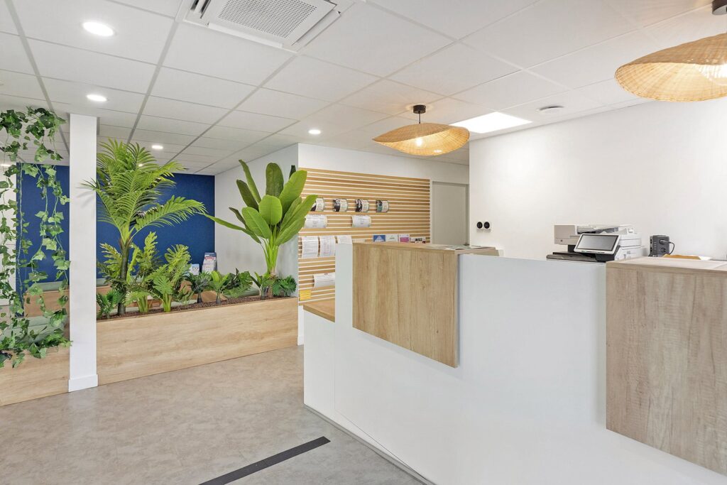 Espace de réception moderne avec un bureau blanc et bois, des plantes d'intérieur, un présentoir à brochures et des lumières au plafond. L'espace présente un design épuré et minimaliste.