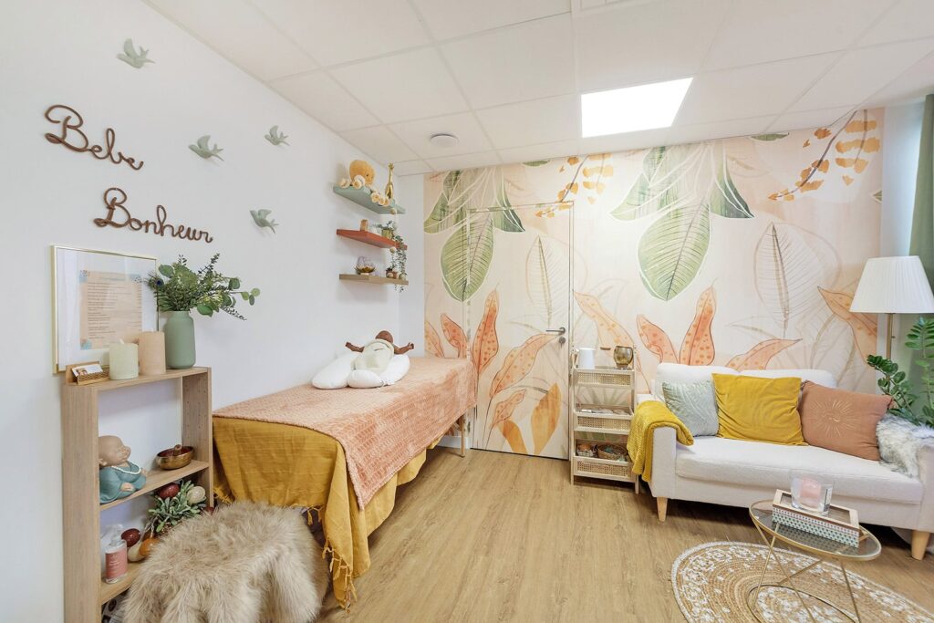 Une chambre confortable avec une table de massage, un canapé, des plantes et un décor pastel. Le mur porte l'inscription "Bébé Bonheur" ; un éclairage doux et un papier peint sur le thème de la nature créent une atmosphère calme.