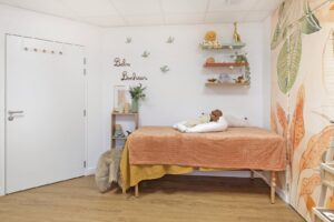 Un lit bien fait avec une literie orange est adossé au mur d'une pièce décorée de plantes, d'étagères et d'un tableau mural où l'on peut lire "Bebe Bonheur". Une poupée et un coussin d'allaitement sont posés sur le lit.