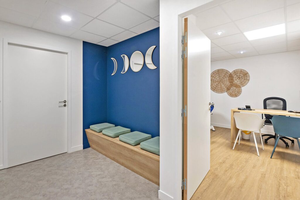 Un intérieur de bureau moderne avec un mur d'accent bleu, des miroirs en forme de croissant de lune, un banc en bois avec des coussins verts, et un espace de bureau adjacent avec un bureau et des chaises.