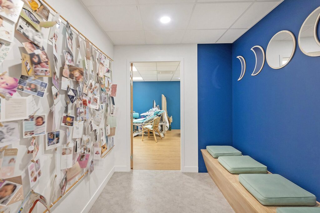 Un couloir avec un tableau d'affichage couvert de cartes et de photos, un banc en bois avec des coussins, et une vue sur une salle avec du matériel médical et des chaises.
