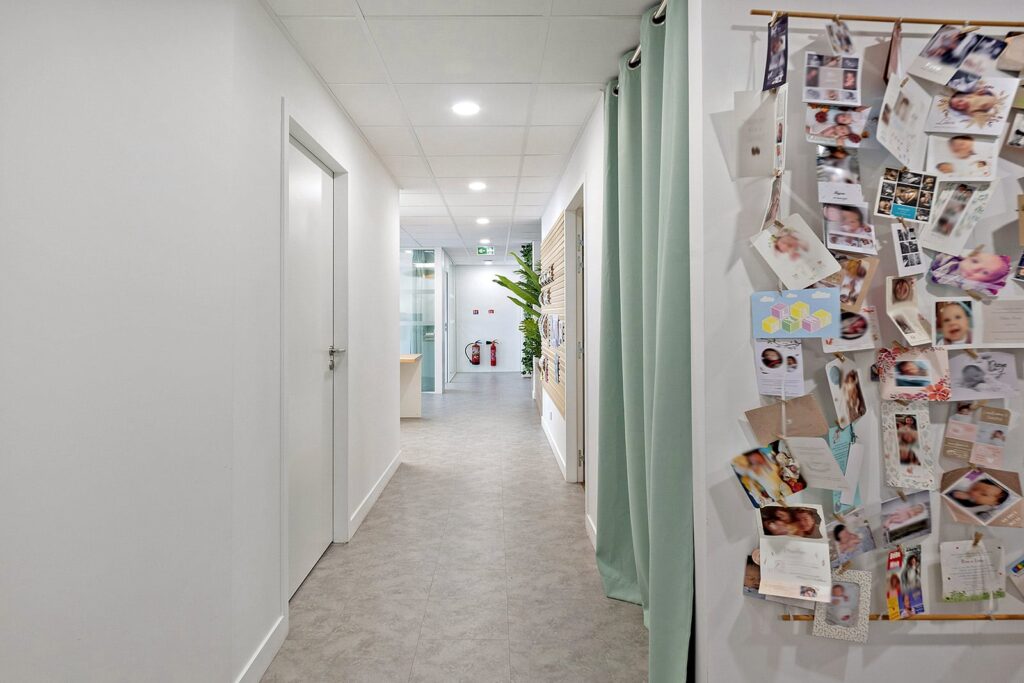 Un couloir avec des portes fermées, des lumières vives au plafond, un tableau d'affichage avec de nombreuses photos de bébés et des cartes, et une plante en pot au fond.