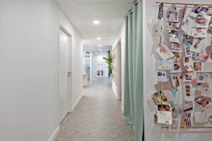 Un couloir avec des portes fermées, des lumières vives au plafond, un tableau d'affichage avec de nombreuses photos de bébés et des cartes, et une plante en pot au fond.