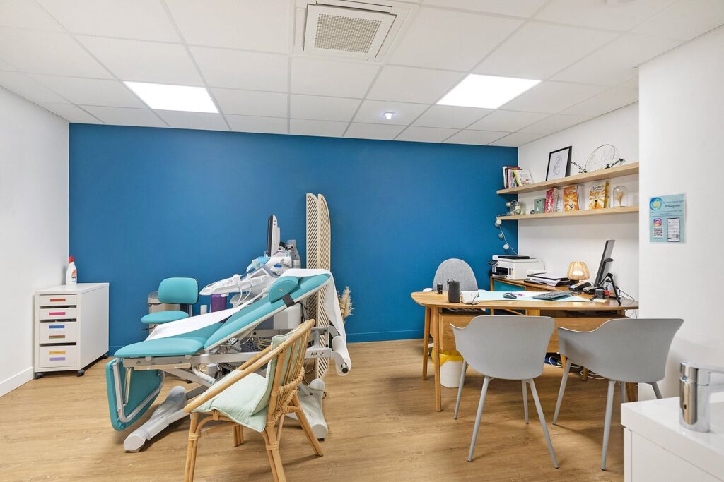 Un cabinet médical moderne avec une chaise d'examen, un bureau, un ordinateur, des étagères avec des modèles médicaux et des chaises pour les patients sur un mur d'accent bleu.