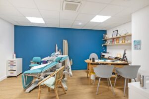 Un cabinet médical moderne avec une chaise d'examen, un bureau, un ordinateur, des étagères avec des modèles médicaux et des chaises pour les patients sur un mur d'accent bleu.