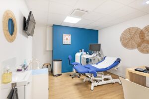 Une salle d'examen médical propre et moderne avec un mur d'accent bleu, une table d'examen, du matériel médical, un ordinateur et des décorations murales.