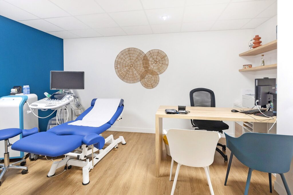 Une salle d'examen médical moderne avec une table d'examen bleue, un appareil à ultrasons, un bureau avec des chaises, un ordinateur et des œuvres d'art murales décoratives.