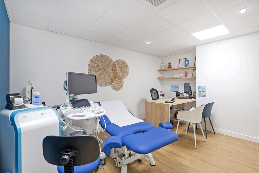 Une salle d'examen médical moderne avec une chaise d'examen bleue, un équipement d'échographie, un bureau avec des chaises et des éléments décoratifs au mur.