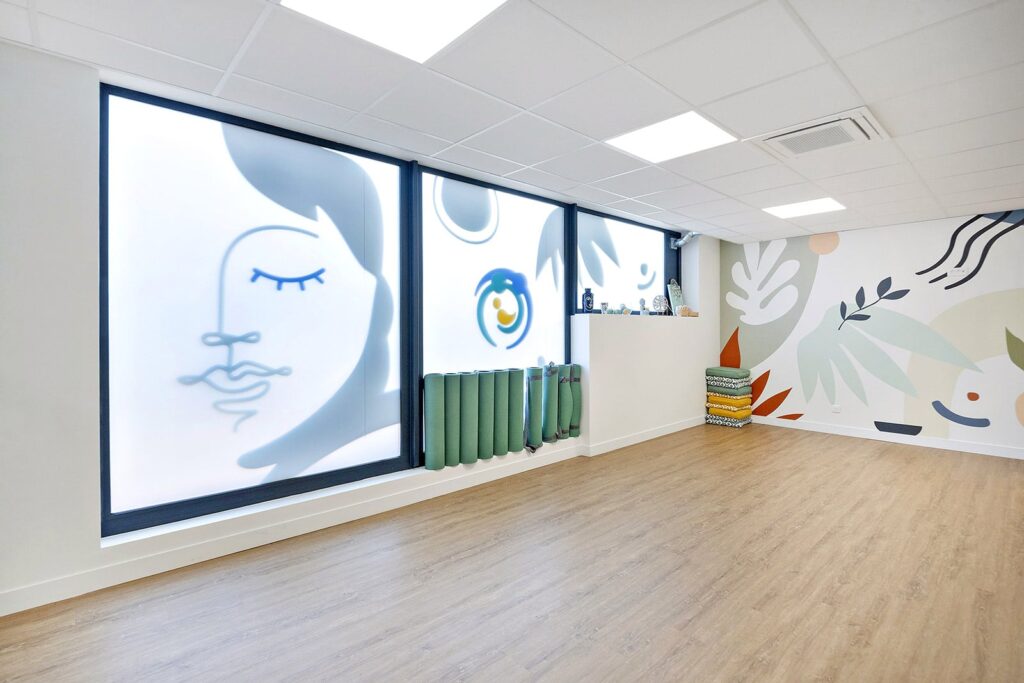 Studio de yoga moderne et lumineux avec sol en bois, art mural abstrait, fenêtres givrées avec illustration de visage, tapis de yoga et couvertures pliées soigneusement empilées sur une étagère.
