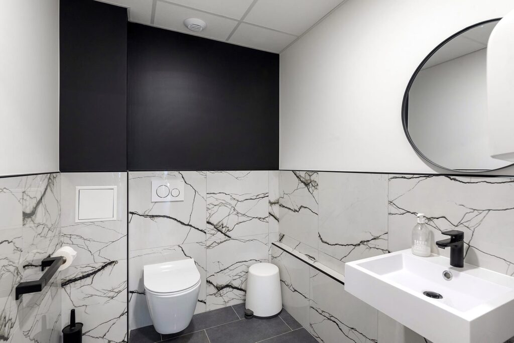 Salle de bains moderne avec toilettes murales, lavabo rectangulaire, miroir rond et murs en marbre blanc. Luminaires noirs et couleurs monochromes.