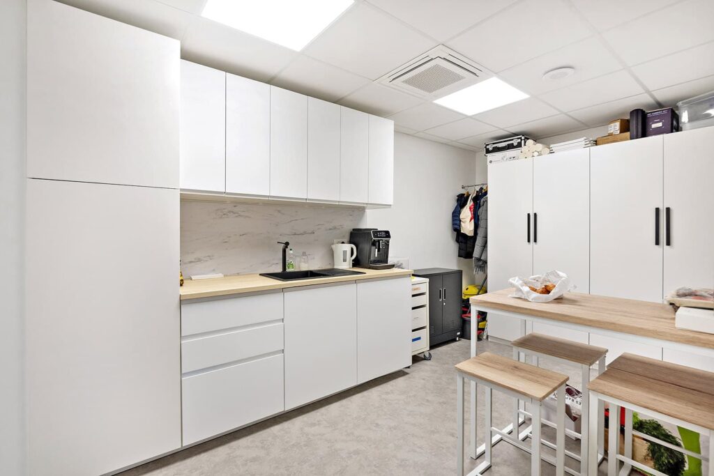 Une cuisine de bureau moderne avec des armoires blanches, un comptoir en bois, un évier, une machine à café, des meubles de rangement, une table haute et des tabourets sous des plafonniers lumineux.