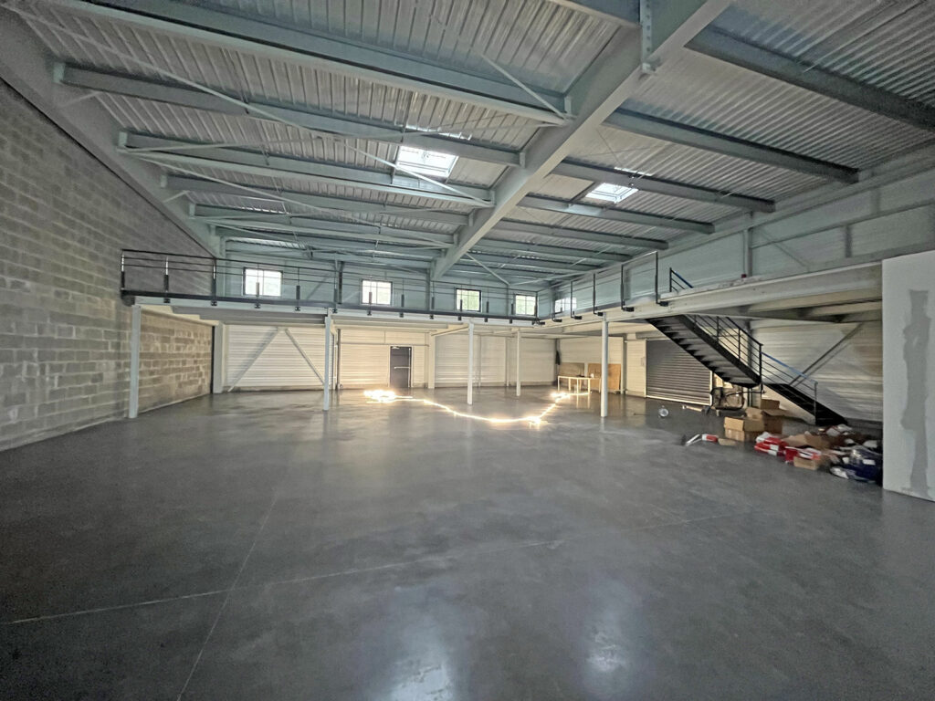 Spacieux entrepôt vide avec sol en béton, haut plafond, mezzanine, escaliers en métal et quelques boîtes empilées dans un coin. La lumière naturelle pénètre par les fenêtres et les puits de lumière.
