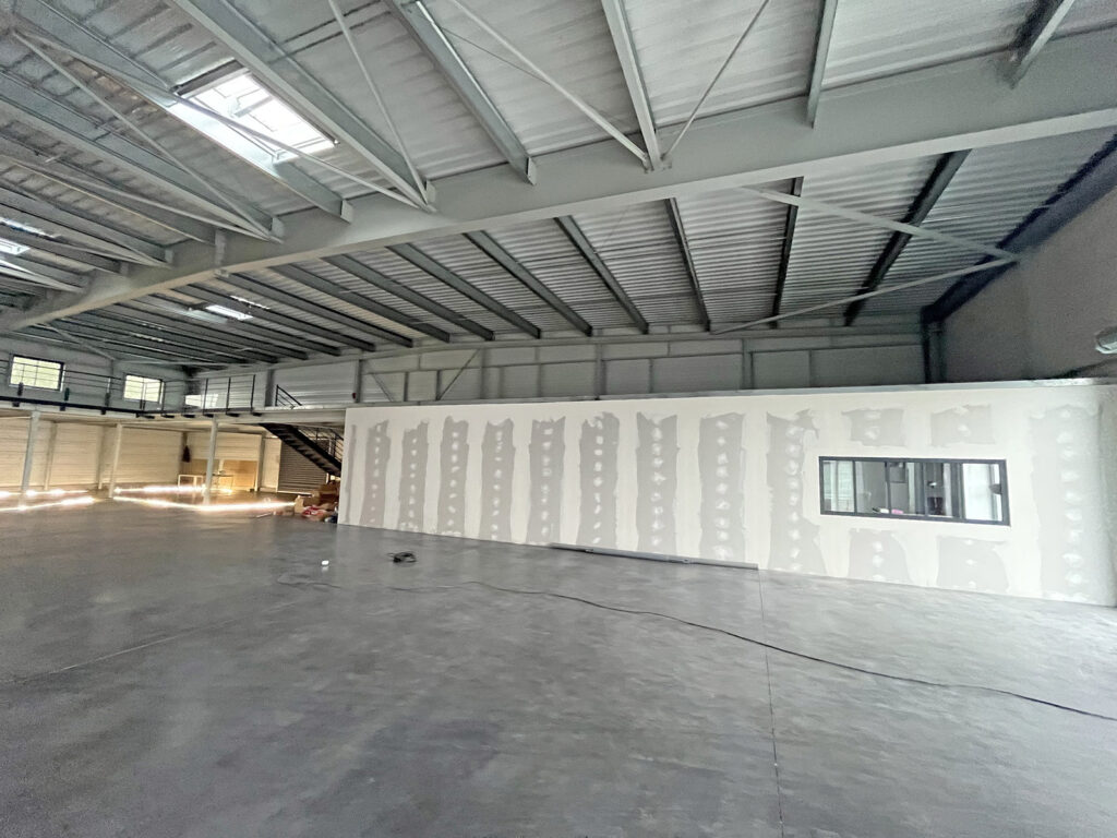 Intérieur d'un entrepôt inachevé avec des poutres métalliques apparentes, une mezzanine, des murs partiellement finis et une seule fenêtre dans une structure blanche.