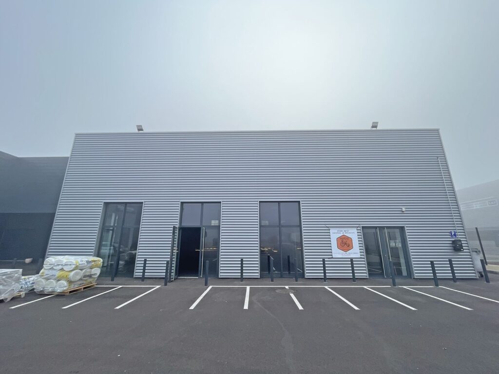 Un bâtiment industriel moderne avec une façade en tôle ondulée, de grandes portes vitrées, une enseigne près de l'entrée, des palettes empilées sur la gauche et un parking vide devant.