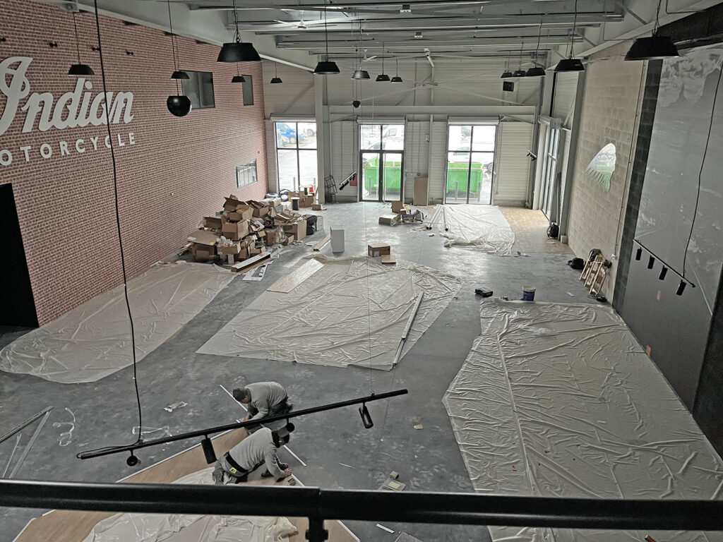 Vue large d'un espace industriel en cours de rénovation, avec des bâches en plastique recouvrant le sol, des boîtes et des fournitures éparpillées, et des ouvriers en train de peindre au premier plan.