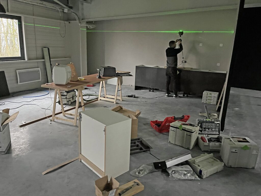 Une personne utilise un niveau laser vert sur un mur dans une pièce en cours de rénovation, avec des outils, des boîtes et des meubles éparpillés dans l'espace inachevé.