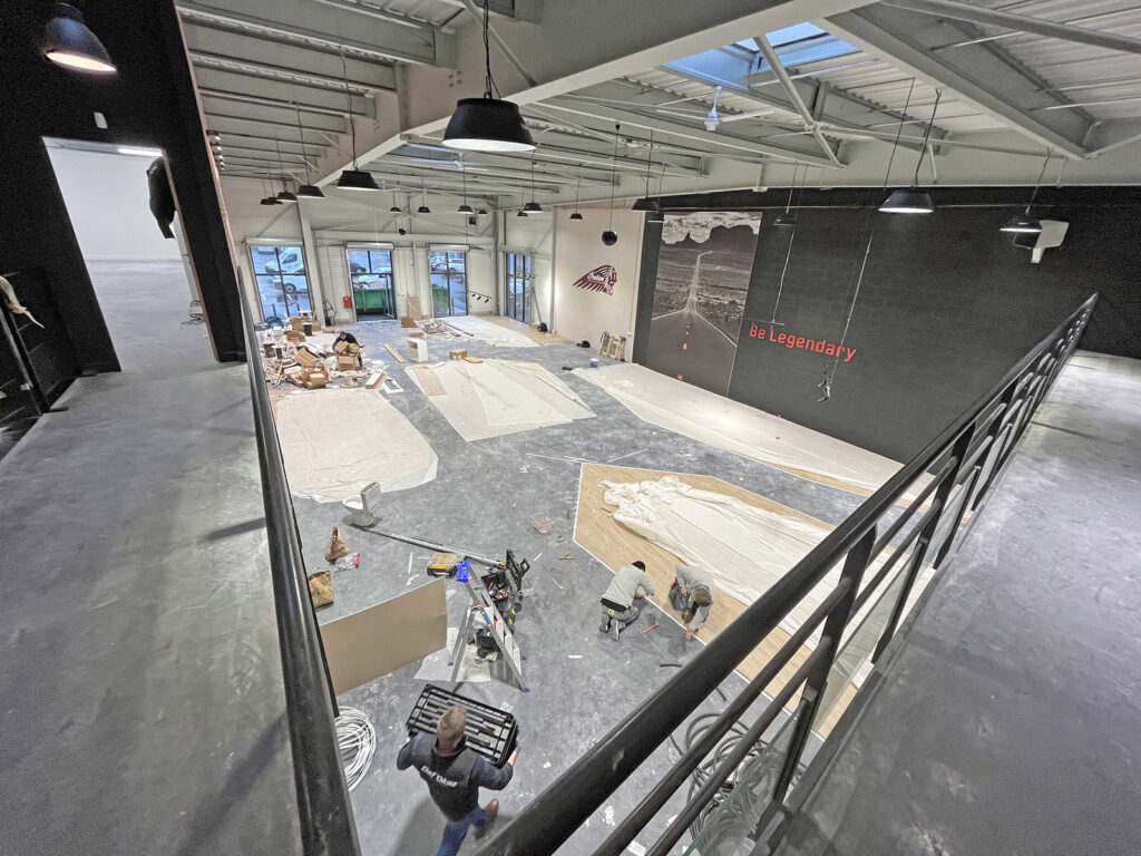 Grand espace intérieur en cours de rénovation avec des outils, des objets couverts, des travailleurs et un mur sur lequel on peut lire "be Legendary" ; fenêtres et plafonniers visibles.