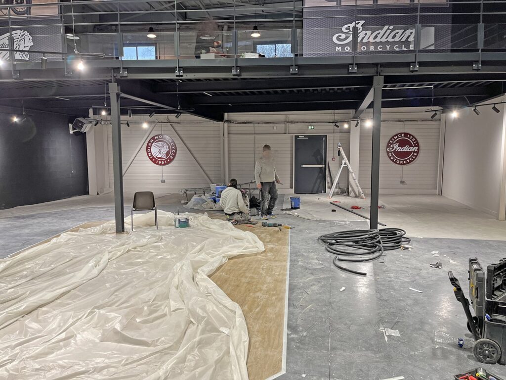 Deux ouvriers sont en train de peindre et de préparer l'intérieur d'une grande salle d'exposition dont les murs sont ornés de logos Indian Motorcycle ; des matériaux de construction et des équipements sont éparpillés autour.
