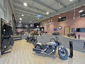Intérieur d'une salle d'exposition avec plusieurs motos exposées, un panneau "Indian Motorcycle" sur un mur de briques et des étagères de matériel d'équitation sur le côté gauche.