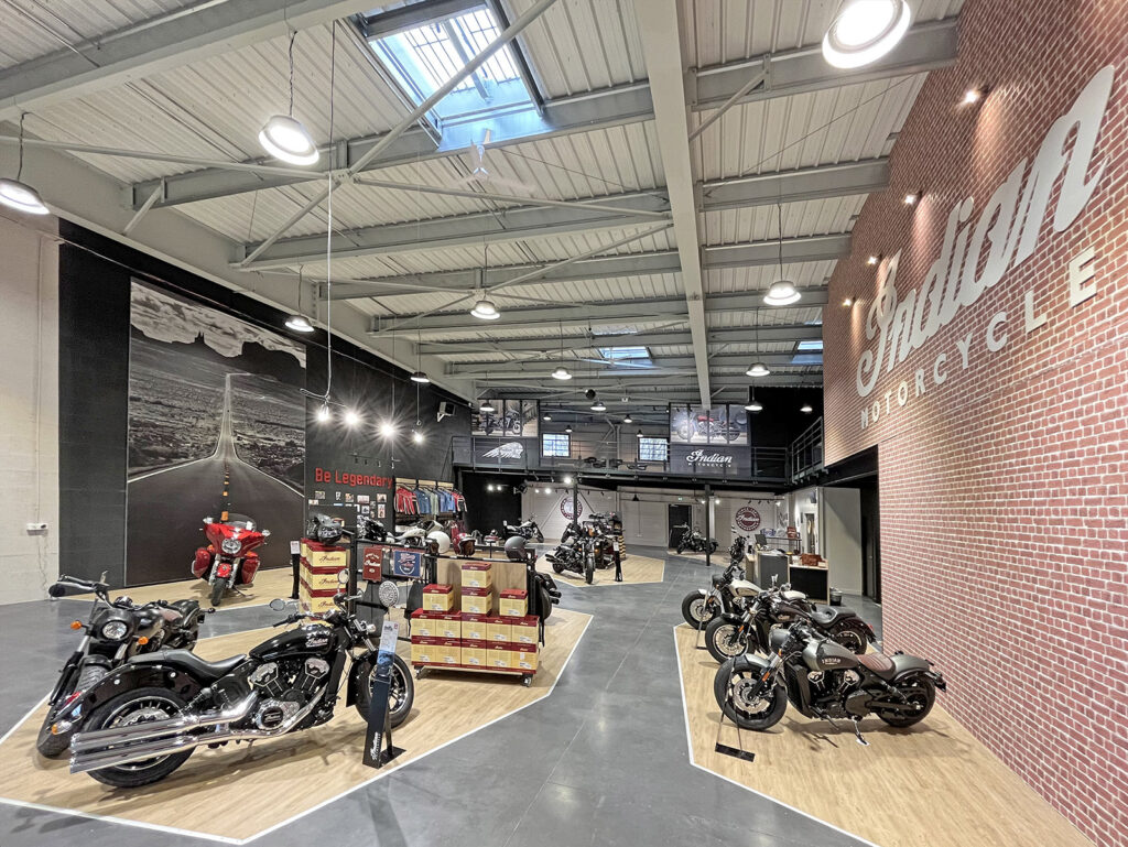 Une salle d'exposition de motos spacieuse avec plusieurs Indian Motorcycles en exposition, de grandes peintures murales, des caisses empilées et un éclairage zénithal lumineux.
