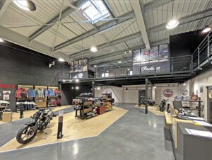 Salle d'exposition de motos moderne avec des motos exposées, des porte-vêtements, un bureau d'accueil, un espace en hauteur, un éclairage et un décor de style industriel.