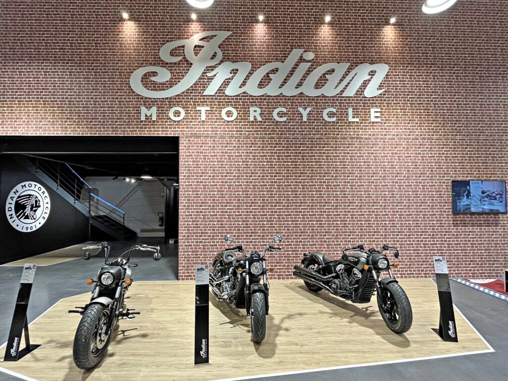 Trois motos indiennes sont exposées sur une plate-forme devant un mur de briques surmonté d'un grand panneau "Indian Motorcycle".