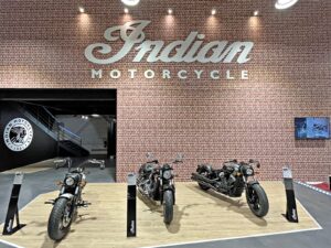 Trois motos indiennes sont exposées sur une plate-forme devant un mur de briques surmonté d'un grand panneau "Indian Motorcycle".