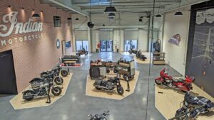 Vue large d'une salle d'exposition de motos avec plusieurs Indian Motorcycles en exposition, des enseignes de marque sur le mur, et des vêtements et accessoires au centre.