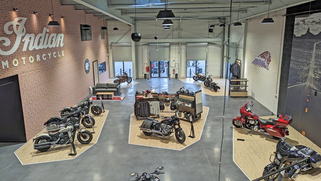 Intérieur d'une salle d'exposition de motos présentant plusieurs Indian Motorcycles, avec des accessoires, des porte-vêtements et des sièges disposés sur des plates-formes en bois.