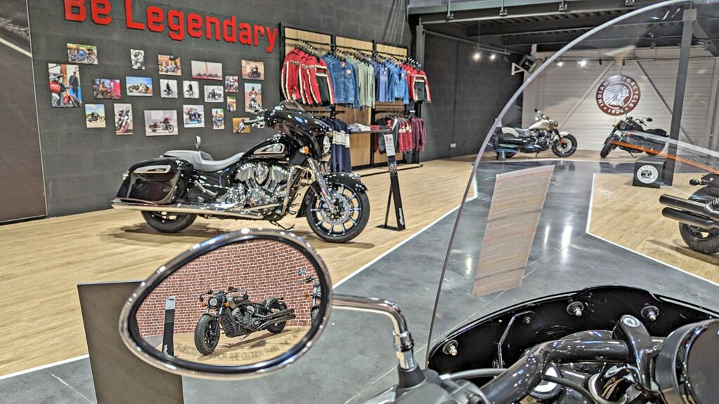 Une salle d'exposition de motos présente plusieurs motos, du matériel de pilotage et des vêtements sur des étagères, une moto se reflétant dans un rétroviseur latéral au premier plan.