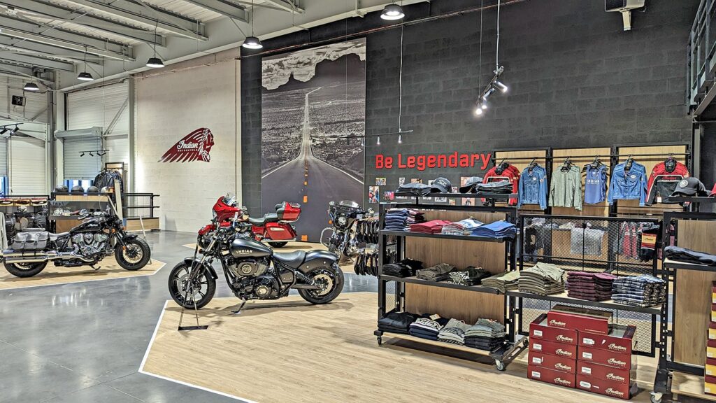 Salle d'exposition de motos avec des motos exposées, des vêtements et des accessoires sur des étagères, ainsi qu'une grande peinture murale et un panneau "Be Legendary" sur le mur.