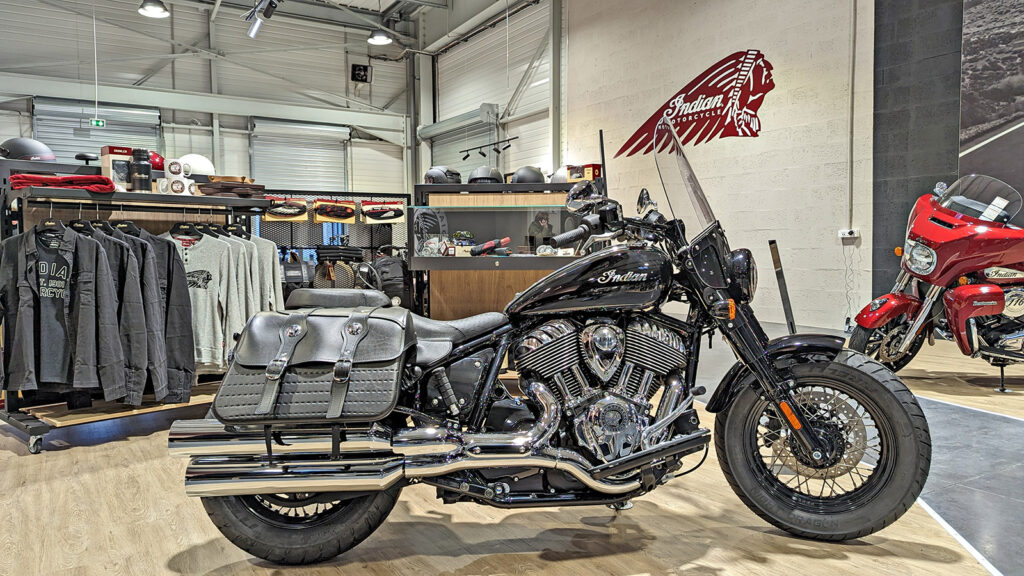 Une moto Indian noire avec sacoches est exposée à l'intérieur d'une salle d'exposition, avec du matériel et des accessoires d'équitation sur des étagères et des rayonnages à l'arrière-plan.